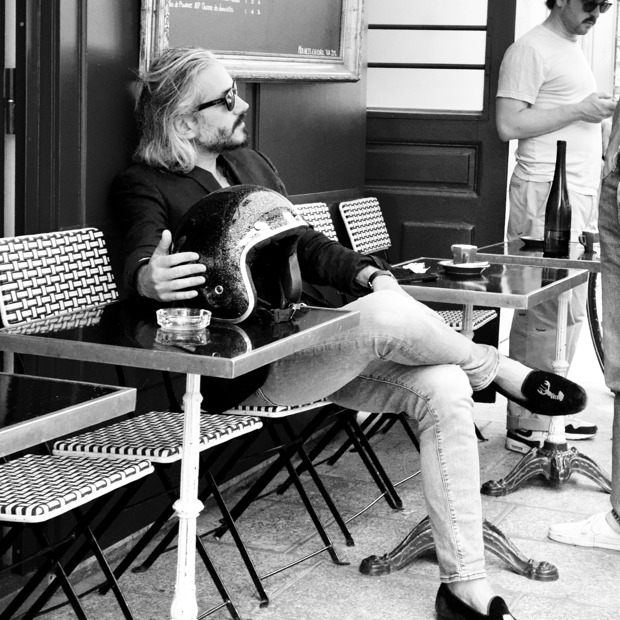 A black and white photo of a man sat outside a cafe with his motorcycle helmet on a table