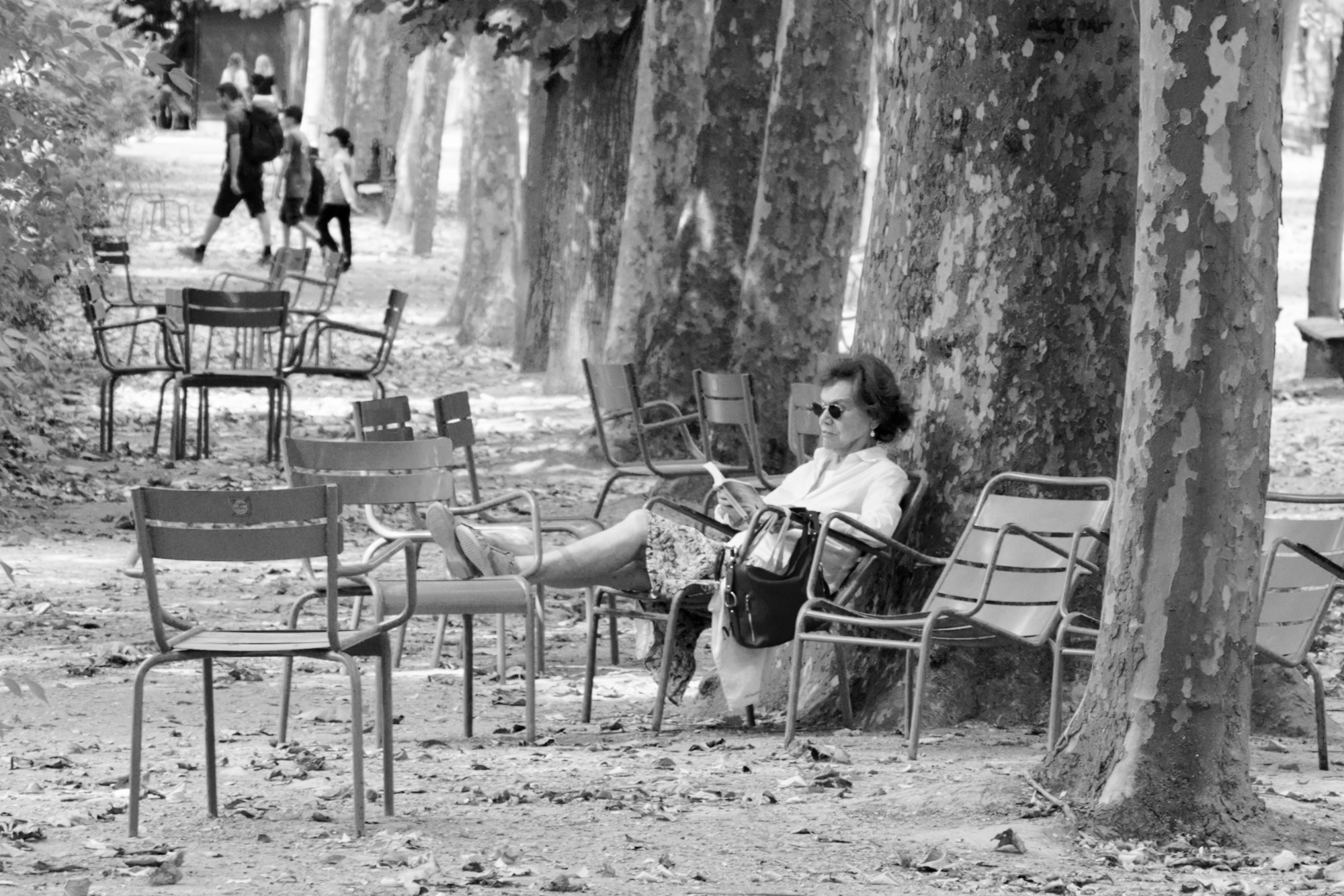 A black and white photo of a woman surrounded by chairs sat under a tree